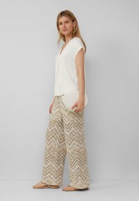 Witte, korte mouwen top met een v-hals, gedragen met brede, beige broek met zigzagpatroon. Houdt een ronde, witte clutch vast.