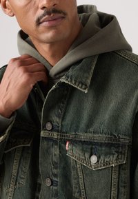 Πράσινο τζιν μπουφάν με ασημένι α κουμπιά, τσέπες στο στήθος και έντονη ραφή. Φορεμένο πάνω από γκρι φούτερ με εμφανές κορδόνι στο καπό.