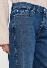 Μπλε τζιν παντελόνια με μεταλλικά καρφιά κατά μήκος της πλευρικής ραφής. Το ύφασμα έχει μια ελαφρώς φθαρμένη υφή με σχεδιασμό κανονικού μήκους και ίσιες γραμμές.