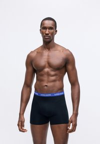 Muscular man wearing black boxer briefs with a blue waistband reading "BJÖRN BORG," standing against a plain light background.