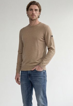 Homme aux cheveux châtain clair portant un pull beige à manches longues et un jean bleu, debout avec une main dans la poche sur un fond uni.