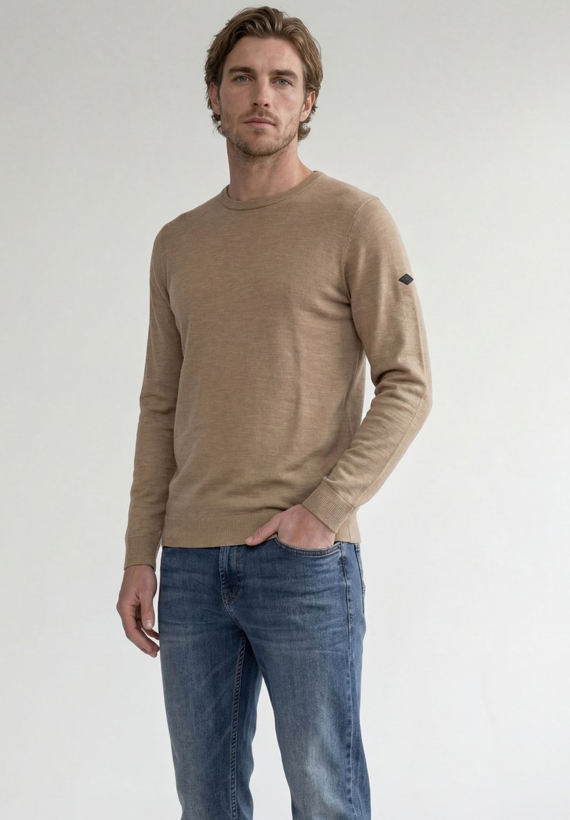 Homme aux cheveux châtain clair portant un pull beige à manches longues et un jean bleu, debout avec une main dans la poche sur un fond uni.