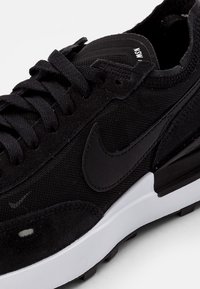 Baskets noires avec tige en mesh et en daim, lacets noirs, semelle intermédiaire blanche et semelle extérieure texturée, avec un logo Swoosh ton sur ton sur le côté.