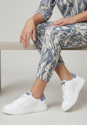 Osoba sedící na lavičce, oblečená do modro-béžových vzorovaných kalhot a bílých tenisek se šedými akcenty, ukazující uvolněnou pózu a zkřížené nohy.