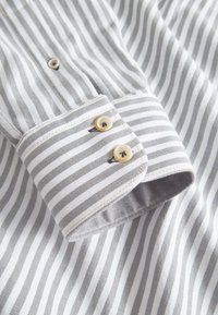 Colletto di una camicia a righe grigie e bianche con polsini arrotondati, dotato di due bottoni beige e un dettaglio di cucitura a contrasto su un tessuto liscio.