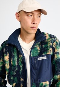 Columbia fleecejacka med ett flerfärgat mönster i grönt, beige och marinblått, med en dragkedjeförsedd bröstficka och hög krage.