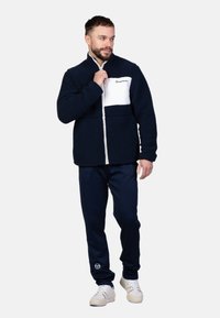 Marineblaue Fleecejacke mit weißem Brustpanel, Reißverschluss und hohem Kragen; kombiniert mit marineblauen Hosen und weißen Sneakern.