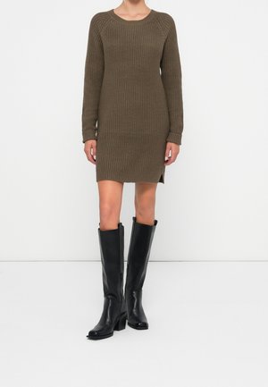 Femme portant des bottes en cuir noires montant jusqu'aux genoux et une robe-pull tricotée vert olive à manches longues sur un fond blanc uni.