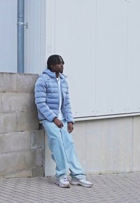 Lys blå pufferjakke, hvid t-shirt og lyse denimjeans. Sneakers i grå nuancer. Betonvæg og grå bygning i baggrunden.