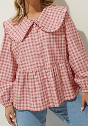 Blusa de cuadros rosa y blanca con mangas largas abullonadas, cuello grande con volantes, cierre de botones en la parte delantera y cintura fruncida, llevada con vaqueros azules.