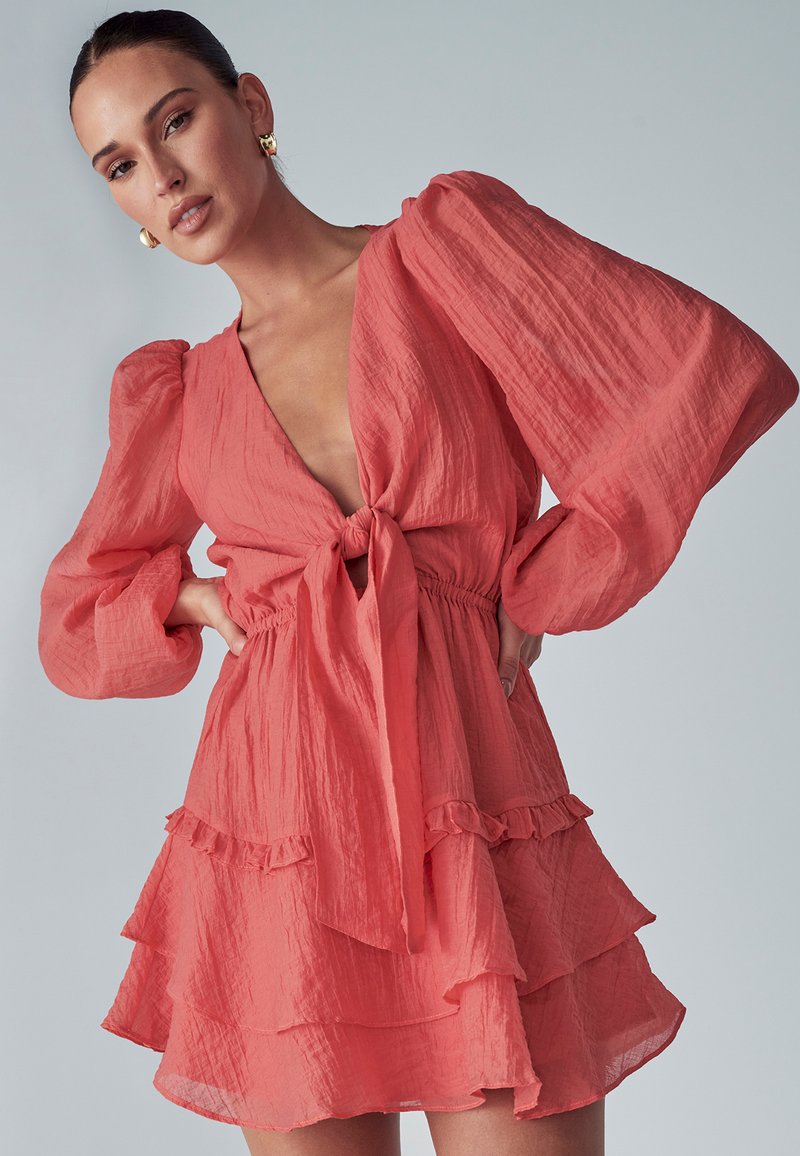 Frau in einem korallenrosa Kleid mit puffartigen langen Ärmeln, einem tiefen V-Ausschnitt, der an der Taille gebunden ist, und einem geschichteten Rüschensaum.