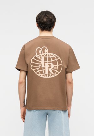 Rövid, göndör hajú személy barna pólót visel, melyen nagy, bézs színű grafika található: egy földgömb, rajzfilmszerű szemek, kéz és a "LR ab" felirat a hátoldalon.