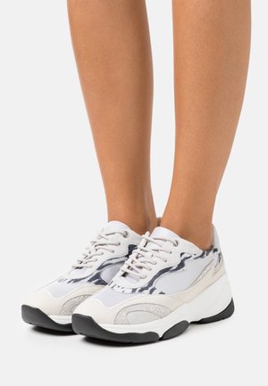 Ben iført hvide og grå chunky sneakers med sorte bølgede mønstre og tykke sorte såler på en hvid baggrund.