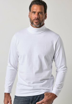 Hombre con barba que lleva una camiseta blanca de cuello alto y manga larga y vaqueros oscuros, de pie frente a un fondo liso y claro.
