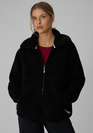 Jeune femme aux cheveux blonds portant une veste polaire noire zippée sur un t-shirt rouge, debout les mains dans les poches devant un fond uni.