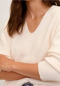 Jasno beżowy sweter dzianinowy z dekoltem w serek, o prążkowanej strukturze i szerokich rękawach, noszony z założonymi na siebie rękoma, prezentujący luźny styl.
