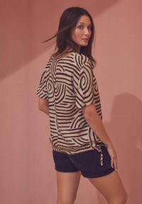 Losse top met een crèmekleurige basis en donkerbruine spiraalpatronen, korte mouwen en een strikdetail aan de zijkant, gecombineerd met donkere shorts.