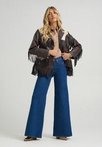 Smeđa kožna jakna s resicama i kremastim detaljima, uparena s plavim zvonastim trapericama. Model stoji prekriženih ruku, prikazujući odjeću.