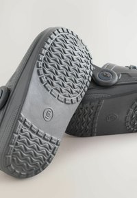 Des chaussures grises à enfiler avec une semelle en caoutchouc texturé affichant des motifs ondulés. Elles sont dotées d'un bouton rond sur le côté pour un enfilage facile.