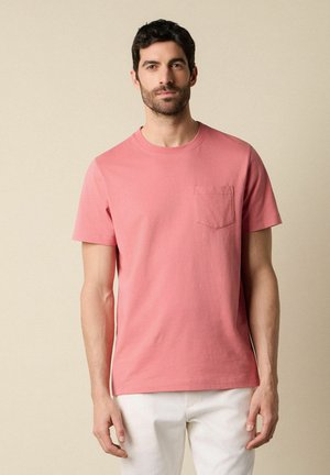 Man met kort donker haar en baard, draagt een zalmroze T-shirt met korte mouwen en borstzak en witte broek tegen een beige achtergrond.