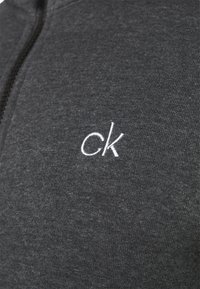 Tmavě šedá zipová mikina vyrobená z měkkého materiálu, s bílou vyšitou značkou "ck" na levé straně. Hladká textura a štíhlý střih.