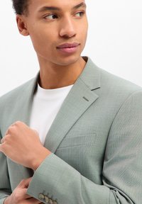 Giovane uomo che aggiusta la manica di una giacca da abito grigio chiaro sopra una camicia bianca, guardando di lato contro uno sfondo bianco semplice.