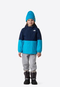 Geacă impermeabilă albastru și bleumarin cu glugă, pantaloni de trening gri și cizme de iarnă negre. Dispune de manșete elastice și un logo pe geacă.