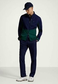 Uomo che indossa una giacca con zip blu navy e verde, pantaloni blu navy, scarpe bianche e un cappellino da baseball blu navy, in piedi con le mani nelle tasche, con lo sguardo rivolto verso il basso.