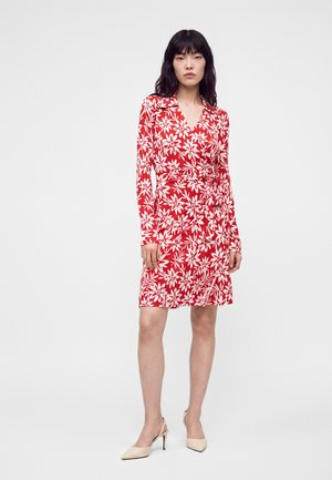 Kvinde med sort hår iført en rød omslagskjole med hvidt blomsterprint og beige højhælede sko, stående mod en hvid baggrund.