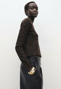 Top de manga larga con estampado de leopardo en marrón y negro, combinado con una falda de cuero negra. Modelo llevando una pulsera de brazalete con un diseño llamativo en dorado.