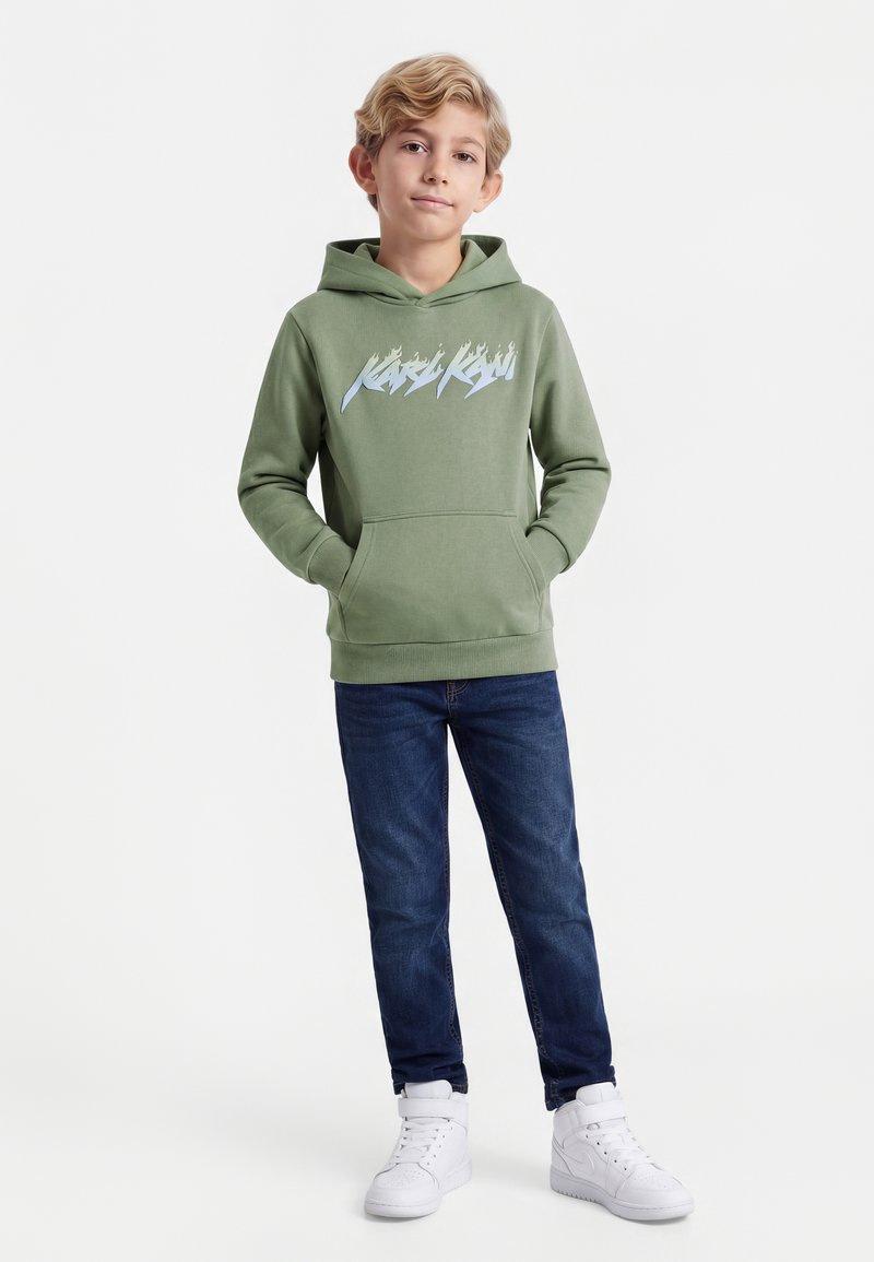Chłopiec o krótkich blond włosach, ubrany w zieloną bluzę z kapturem z napisem, niebieskie dżinsy i białe trampki, stoi z rękami w kieszeniach.