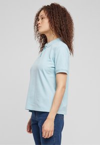Lys blå polo skjorte med krage, korte ermer og avslappet passform. Brukt med mørkeblå jeans, sett fra siden mot en ensfarget bakgrunn.