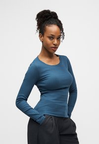 Νεαρή γυναίκα με κυματιστά μαλλιά σε κότσο, φοράει μπλε ριμπ μπλούζα με μακριά μανίκια και σκουρόχρωμο παντελόνι με τα χέρια στις τσέπες, στέκεται μπροστά σε απλό φόντο.