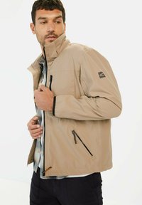 Beige ritsjack met een staande kraag, twee zijzakken, zwarte ritsen, textuurstof en logoaccent op de mouw.