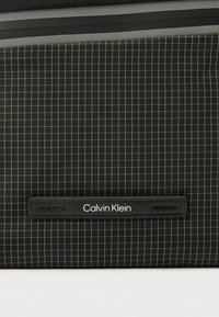 Fekete és szürke kockás szövet, középen varrott, fekete bőr téglalap alakú címkével, amelyen fehér betűkkel olvasható a „Calvin Klein” felirat.