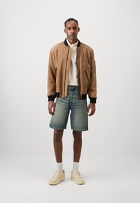 Jachetă bomber din suede maro cu detalii negre, cămașă albă cu mânecă lungă, pantaloni scurți din denim albastru și adidași crem. Textură netedă, croială lejeră.