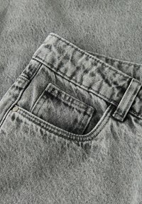 Nærbilde av falmet grå denimjeans med fokus på en fremre lomme med synlige sømmer og en liten metallnagle.