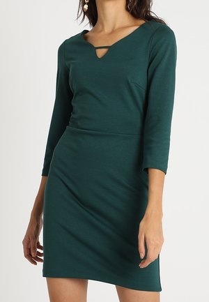 Frau trägt ein figurbetontes dunkelgrünes Kleid mit Dreiviertelärmeln und einem kleinen V-förmigen Ausschnitt am Halsausschnitt.