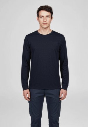 Jonge man staand en naar voren kijkend, gekleed in een effen donkerblauw shirt met lange mouwen en bijpassende cargobroek tegen een effen achtergrond.