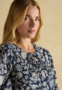 Blonde Frau mit blauen Augen trägt eine marineblaue Bluse mit floralen Mustern und blickt in die Kamera, vor einem sanften, neutralen Hintergrund.