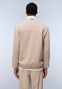 Sudadera beige con una textura suave, puños acanalados y un detalle de costura en la parte posterior. Lleva una capa blanca debajo y se combina con pantalones beige a juego.