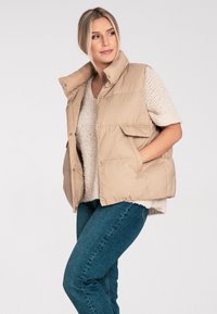 Beżowa kamizelka z wypełnieniem, z wysokim kołnierzem, zapięciem na zatrzaski i dwoma kieszeniami po bokach, noszona na lekkim swetrze i niebieskich dżinsach.