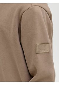 Sudadera beige con una textura suave. Cuenta con un parche de logo en la manga, cuello redondo y un corte relajado. Diseño minimalista.