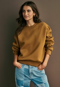 Felpa gialla senape con scollo rotondo e polsini elasticizzati, abbinata a jeans in denim chiaro con toppe, caratterizzati da quadrati di denim a contrasto.