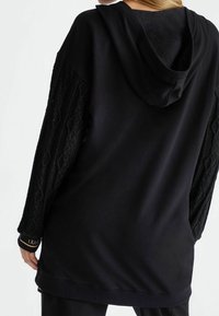 Schwarzer Kapuzenpullover mit strukturierten, gemusterten Ärmeln, ausgestattet mit gerippten Bündchen und einem weichen Baumwollmix. Einfaches Design mit minimalen Akzenten.