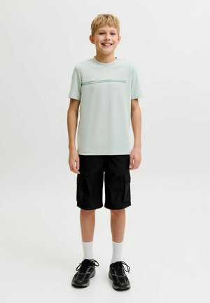 Smilende dreng stående iført lysegrå t-shirt med vandret stribe, sorte cargoshorts, hvide sokker og sorte sneakers på ensfarvet baggrund.