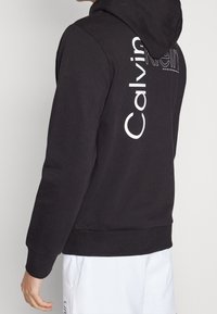 Zwarte hoodie van katoen met een gestructureerde afwerking. Voorzien van het witte Calvin Klein-logo dat verticaal op de achterkant is gedrukt.