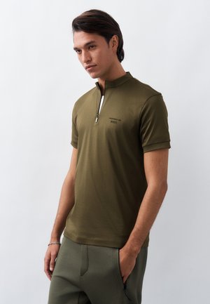 Νέος άνδρας με σκούρα μαλλιά, ντυμένος με κοντομάνικο πόλο πουκάμισο ελιάς με φερμουάρ και αντίστοιχο παντελόνι, που στέκεται μπροστά σε απλό φωτεινό φόντο.