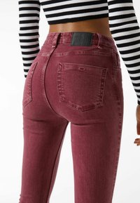 Burgundy tight jeans med hög midja, fem fickor och en texturerad yta. Jeansen har en svart läderetikett i midjan bak.