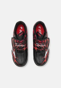 Zapatillas de fútbol negras con patrones abstractos en rojo y blanco, que cuentan con una correa de velcro y un forro acolchado rojo para mayor comodidad.
